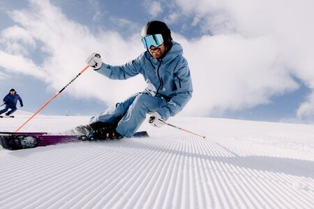Un homme abillé en bleu, portant un casque et un masque, en pleine action qui descend une pente de ski. Le paysage est ensoleillé, ave c quelques nuages. 