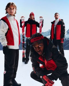 3 personnes debout dont un avec une paire de ski dans les mains, et une personne accroupi en premier plan dans un paysage enneigé